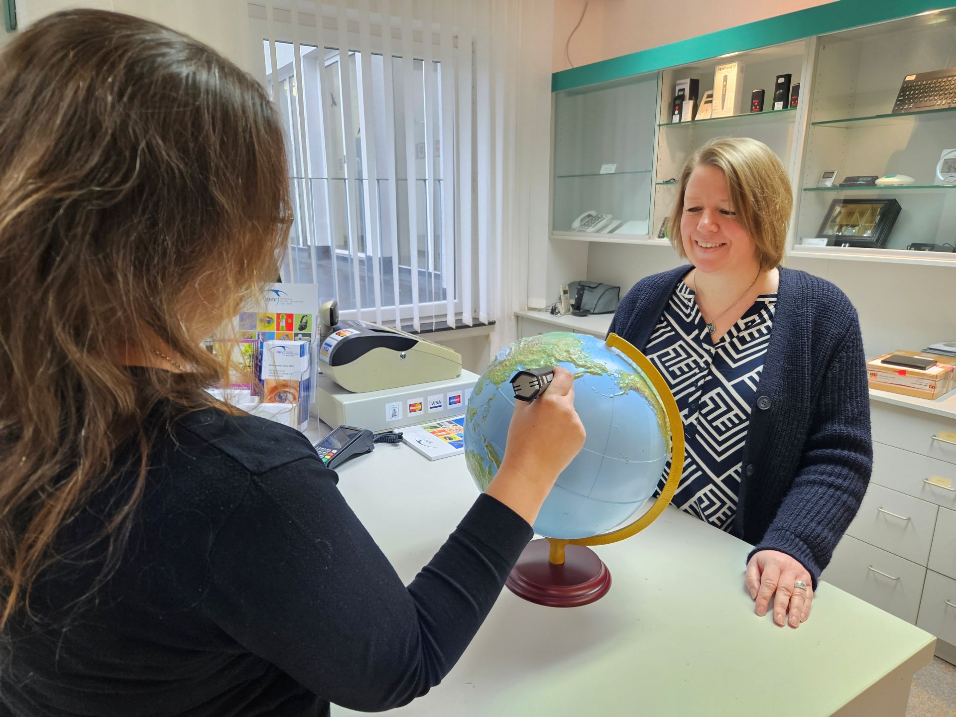 An einem Ladentresen probiert eine Frau mit einem Stift einen sprechenden Globus aus. Die Frau hinterm Tresen lächelt freundlich.